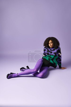 A young woman with vibrant purple tights and sweater sits gracefully on the ground against a matching background in a studio.