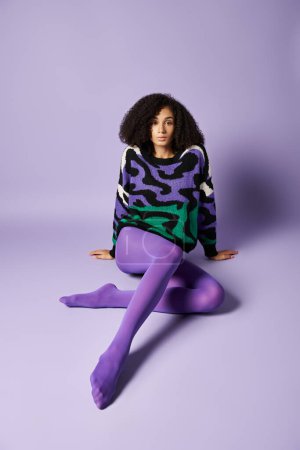 Foto de Una mujer joven en medias vibrantes y suéter se sienta con las piernas cruzadas sobre un fondo púrpura en una pose serena y relajada. - Imagen libre de derechos