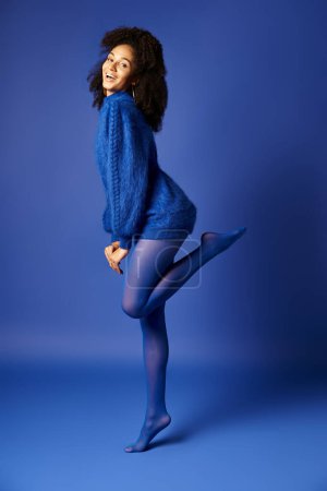 Téléchargez les photos : Une jeune femme élégante en collants vibrants et un pull bleu frappant une pose sur un fond assorti dans un studio. - en image libre de droit