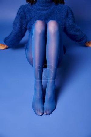 Une jeune femme en robe bleue saisissante pose assise par terre avec un air de sérénité et d'élégance.