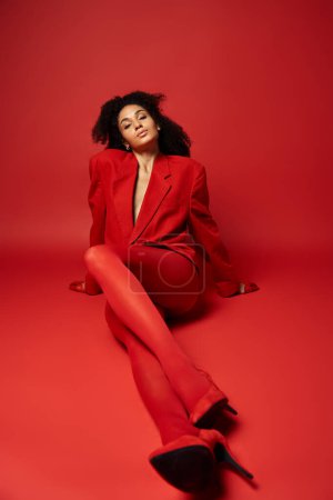 Téléchargez les photos : Une jeune femme en costume rouge s'assoit gracieusement sur le sol rouge vif d'un studio. - en image libre de droit