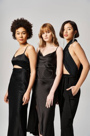 Drei Frauen verschiedener Ethnien stehen in schwarzen Kleidern vor grauem Hintergrund zusammen und zeigen Vielfalt und Eleganz.
