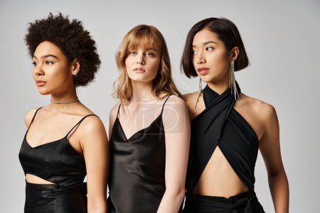 Three women of different ethnicities standing gracefully in black dresses against a neutral grey studio background.
