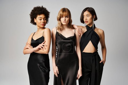 Three women, each in a chic black dress, stand gracefully together against a grey studio background.