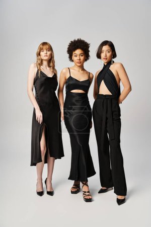 Drei Frauen verschiedener Ethnien stehen in schwarzen Kleidern in einem Studio vor grauem Hintergrund zusammen.