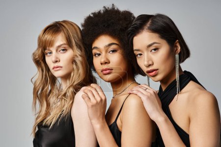 Three diverse women of Caucasian, Asian, and African American descent stand gracefully together against a grey studio background.