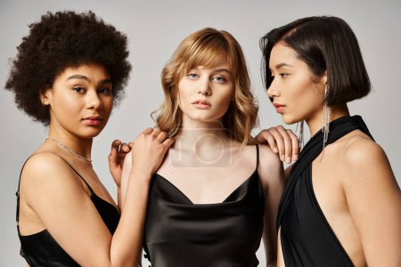 Three beautiful women of Caucasian, Asian, and African American descent standing elegantly on a grey studio background.