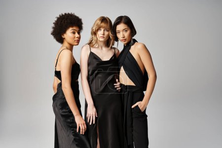 Eine Gruppe von drei schönen Frauen kaukasischer, asiatischer und afroamerikanischer Abstammung, die anmutig vor einer grauen Studiokulisse zusammenstehen.