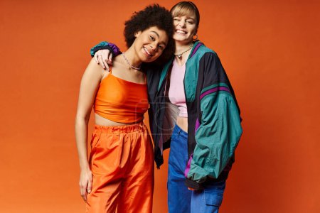 Zwei schöne Frauen unterschiedlicher kultureller Herkunft stehen zusammen in einem Atelier vor orangefarbenem Hintergrund.