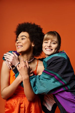 Two women, of different ethnicities, close in a warm hug, smiles radiant against an orange backdrop.