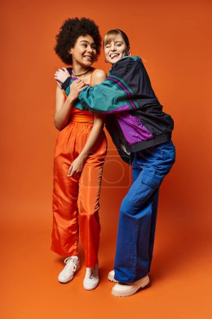 Foto de Una hermosa mujer de pie junto a un amigo en pantalones naranja sobre un fondo naranja vibrante. - Imagen libre de derechos