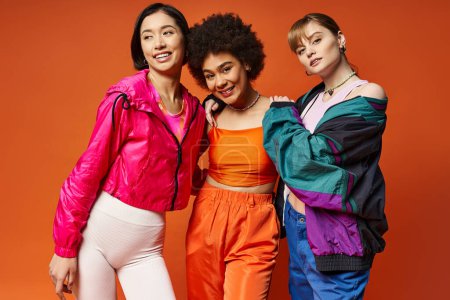 Three women of different ethnicities stand confidently in a studio with an orange backdrop, showcasing multicultural beauty.