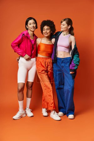 Three women of diverse backgrounds stand united, representing beauty and strength against an orange studio backdrop.