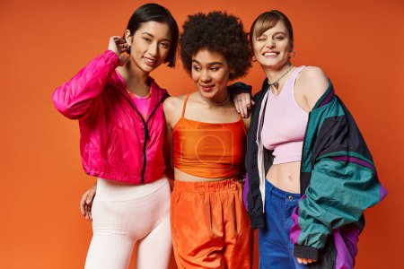 Drei Frauen verschiedener Ethnien stehen gemeinsam vor orangefarbenem Hintergrund und präsentieren multikulturelle Schönheit und Einheit.
