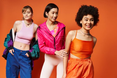 Three women of Caucasian, Asian, and African American descent standing gracefully in a studio with an orange background.
