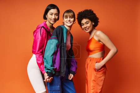 Tres mujeres de diferentes etnias de pie juntas en un estudio sobre un fondo naranja, irradiando belleza y unidad.
