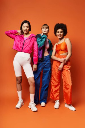 Eine Gruppe schöner Frauen unterschiedlichster Herkunft und ethnischer Zugehörigkeit, die zusammen vor einem orangefarbenen Studiohintergrund stehen.