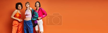 Photo for Three women of different ethnic backgrounds standing together in front of an orange studio background. - Royalty Free Image