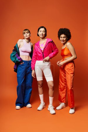Tres hermosas mujeres en un estudio, que representan la diversidad: caucásica, asiática y afroamericana, de pie juntas sobre un fondo naranja.