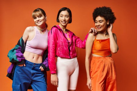 Drei schöne Frauen kaukasischer, asiatischer und afroamerikanischer Abstammung stehen gemeinsam auf einem leuchtend orangefarbenen Studiohintergrund..