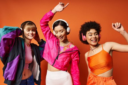 Three diverse women in headphones smiling for a vibrant orange studio photo.