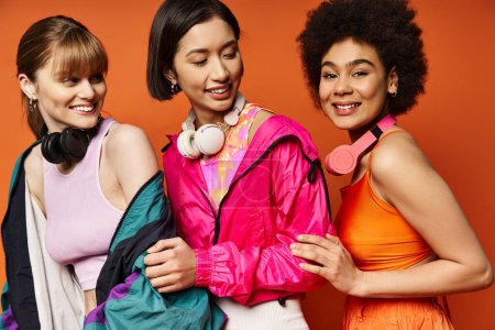 Three women of different ethnicities and backgrounds stand together in a studio against an orange background.