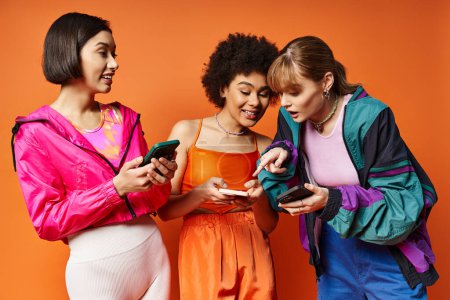 Foto de Tres mujeres diversas con diferentes etnias de pie una al lado de la otra, absorbidas en sus teléfonos celulares contra un fondo naranja. - Imagen libre de derechos
