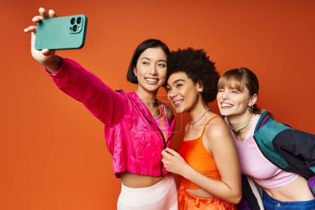 Foto de Tres mujeres, representando diferentes culturas, disfrutando de un momento lúdico mientras se toman una selfie con un teléfono celular sobre un fondo naranja. - Imagen libre de derechos