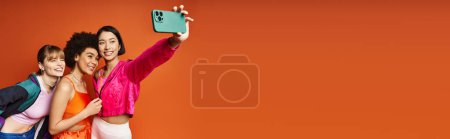 Multikulturelle Frauen machen vor orangefarbenem Studiohintergrund in einem freudigen Moment ein Selfie mit dem Handy.