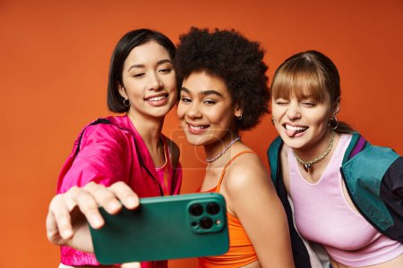 Tres mujeres diversas de ascendencia caucásica, asiática y afroamericana tomando una selfie con un teléfono celular contra un fondo de estudio naranja.