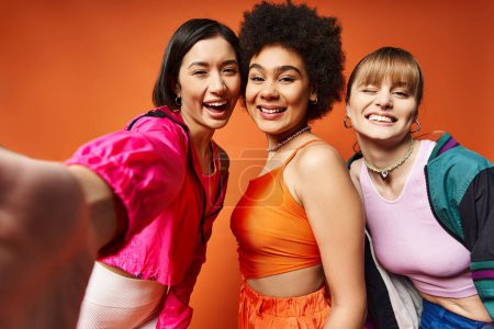 Eine vielfältige Gruppe von Frauen, darunter kaukasische, asiatische und afroamerikanische, stehen vor einer leuchtend orangen Studiokulisse zusammen..