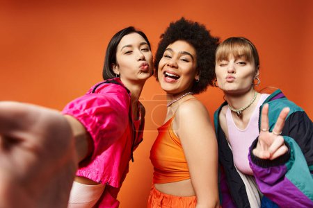 Un grupo de hermosas mujeres jóvenes de diversos orígenes de pie juntos contra un fondo de estudio naranja.