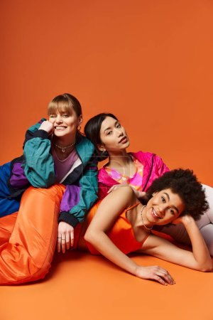 Un groupe diversifié de femmes couchées les unes sur les autres dans une formation pyramidale humaine, sur fond de studio orange.