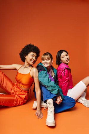 Eine Gruppe schöner junger Frauen verschiedener Ethnien, die eng beieinander in einem Studio vor orangefarbenem Hintergrund sitzen.