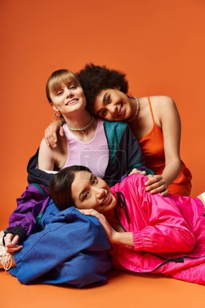 A group of multicultural women, including Caucasian, Asian, and African American, joyfully laying on top of each other on an orange background.