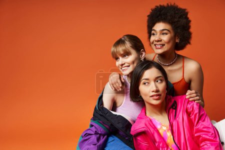 Deux femmes de races différentes posent avec amour avec un enfant heureux devant un fond de studio orange.