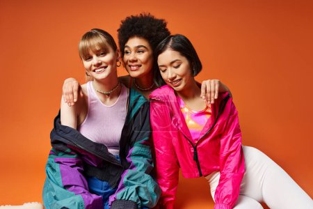 Drei verschiedene Frauen sitzen anmutig auf dem Boden und lächeln für ein Bild, das von einem leuchtend orangefarbenen Hintergrund umrahmt wird..