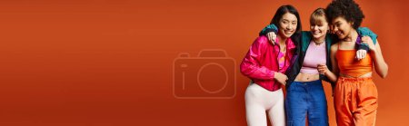 Photo for Three diverse women, Caucasian, Asian, and African American, stand confidently together against an orange studio backdrop. - Royalty Free Image