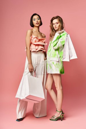 Dos mujeres de diferentes etnias de pie con bolsas de compras sobre un fondo rosa.