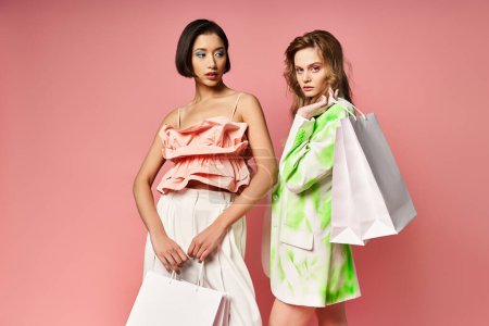 Two women, representing diversity, stand holding shopping bags against a pink studio background.