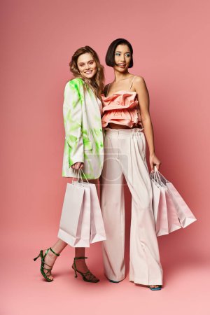 Two women, one Caucasian, one African American, stand with shopping bags against a pink studio background.