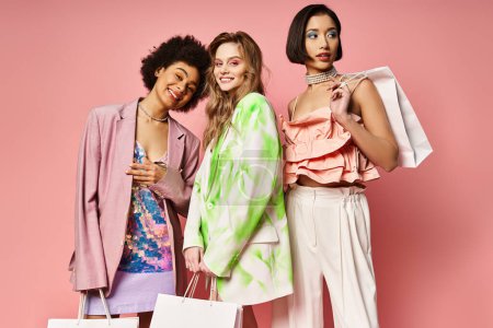 Three women from diverse backgrounds stand together, holding colorful shopping bags in front of a pink studio background.
