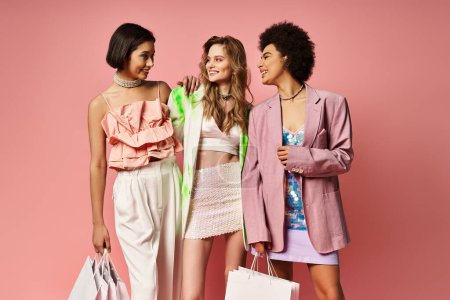 Three women of diverse backgrounds stand together, clutching shopping bags against a pink studio backdrop.
