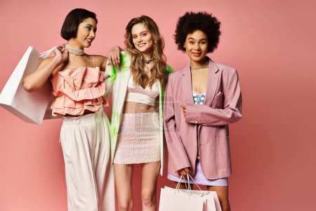 Foto de Tres mujeres de diversos orígenes están de pie lado a lado, sosteniendo bolsas de compras contra un vibrante fondo de estudio rosa. - Imagen libre de derechos