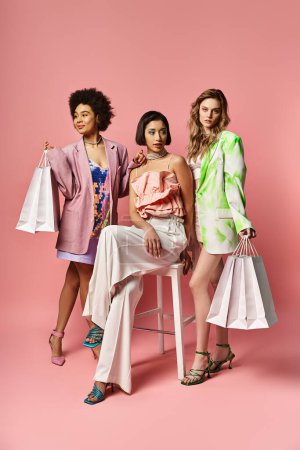Photo for Three diverse women sitting on a stool, holding shopping bags against a pink studio background. - Royalty Free Image