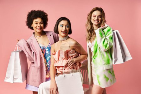 Drei Frauen unterschiedlicher Herkunft posieren elegant mit Einkaufstaschen vor rosa Studiokulisse.