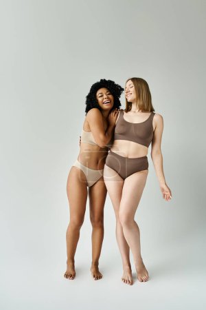 Two beautiful diverse women in cozy pastel underwear stand closely together.