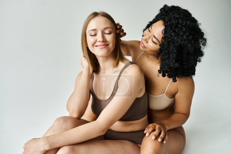 Foto de Dos mujeres diversas en acogedora ropa interior pastel se sientan juntas sobre un fondo blanco. - Imagen libre de derechos