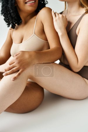 Two beautiful women of different ethnicities sitting closely in pastel underwear.