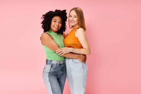 Two attractive diverse women in cozy attire share a heartfelt hug in front of a pink backdrop.